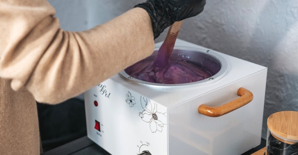 An esthetician wearing gloves stirring purple wax in a wax pot indoors.